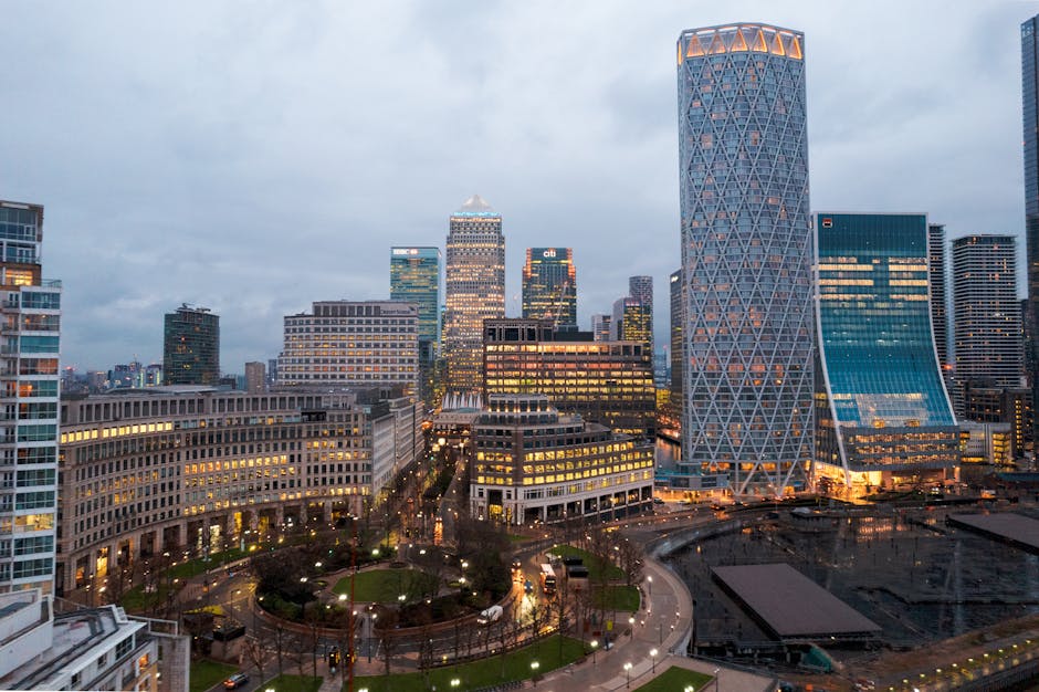 A wide view of the London skyline at sunset, featuring numerous modern high-rise buildings and skyscrapers situated along the riverbank. In the foreground, part of a black metal railing is visible, sl