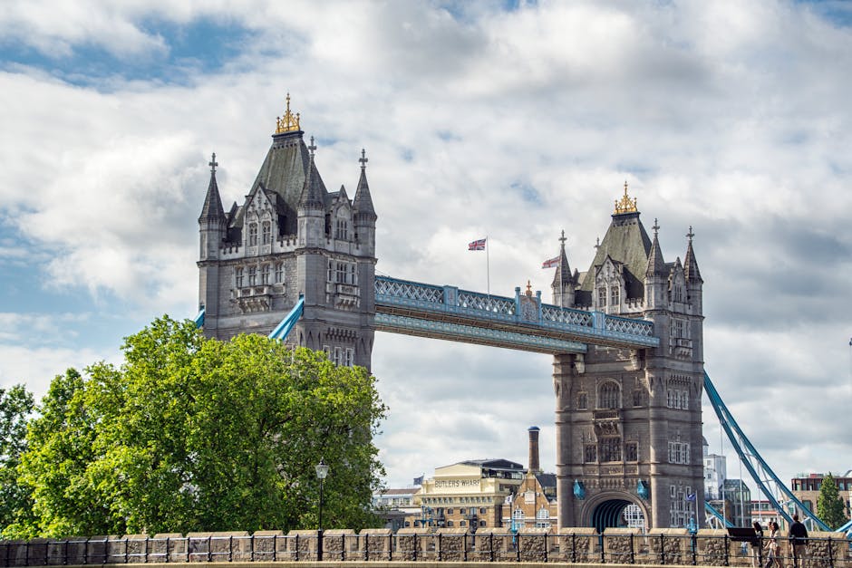 A view of Tower Bridge in London showing the structure's two large towers connected by upper walkways, with a clear blue sky and scattered clouds in the background. The bridge's lower section features