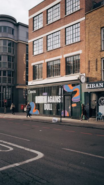 The image shows the exterior of a multi-storey brick building in Shoreditch, with large windows and a storefront advertising an art exhibition titled 'DESIGN AGAINST DULL' displayed prominently on the